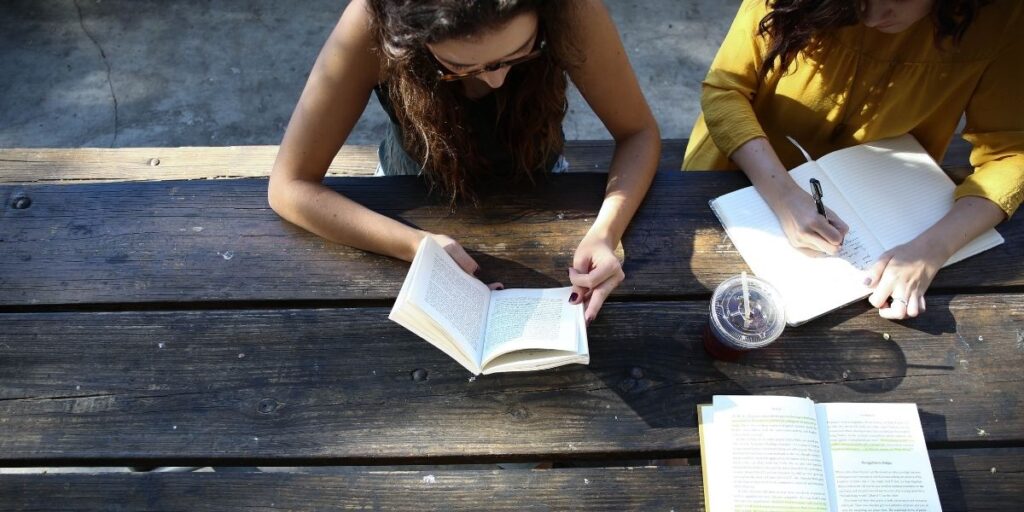 2 women and 1 man sitting on an outside table reading books and studying.