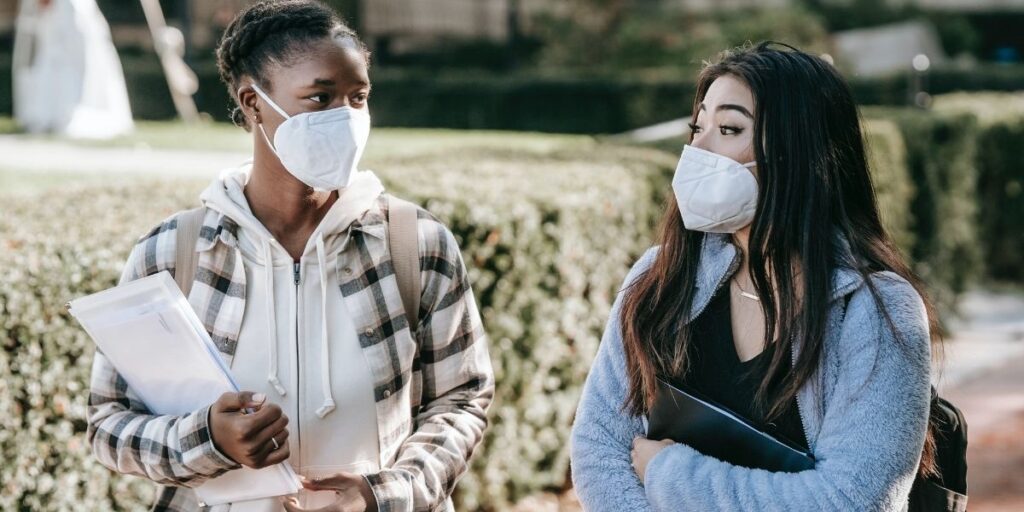 Two women wearing masks, walking and talking, holding a note books and listening to each other