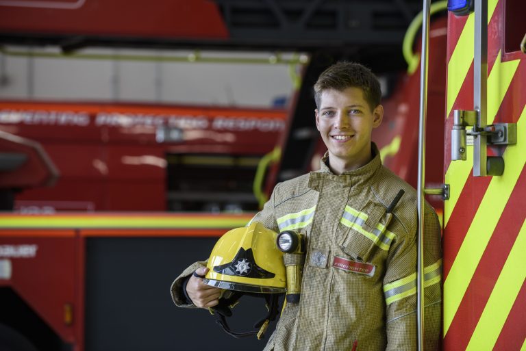 Young adult male firefighter holds his helmet while leaning on a fire engine.