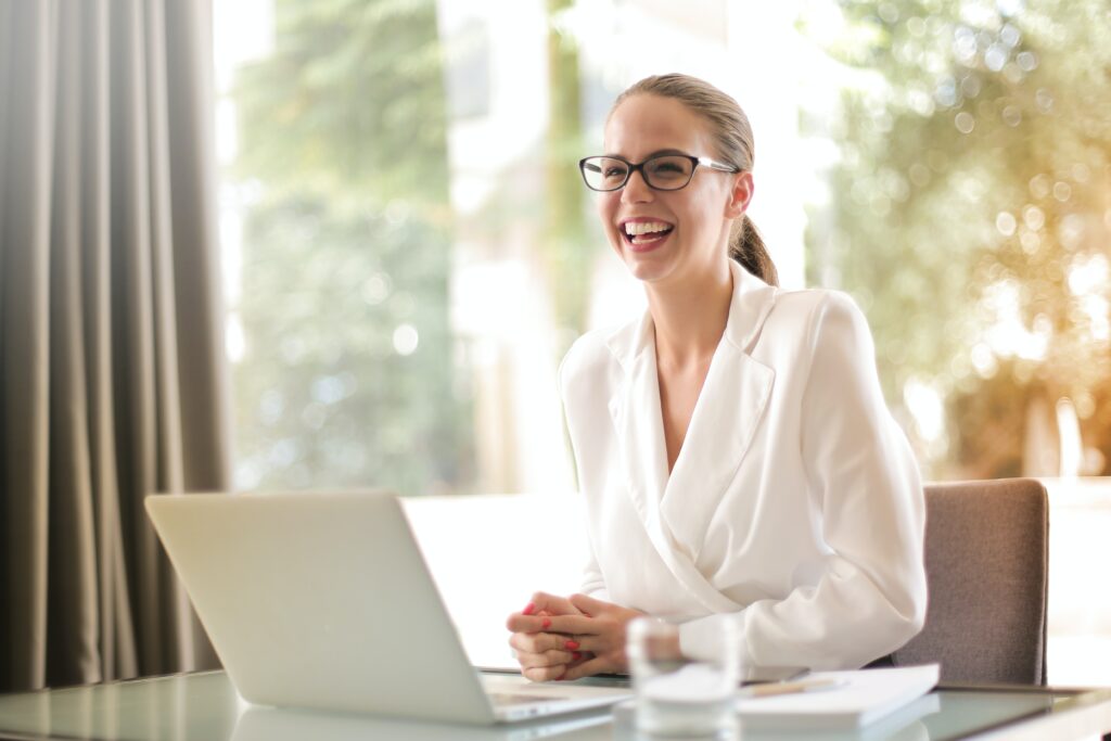 Female woman, sitting at a desk with a silver laptop opened. She is smiling and had black glasses on, blonde hair tied in a pony and has her back facing a large window with sunlight beaming in and trees in the back ground. This woman is working for an apprenticeship.