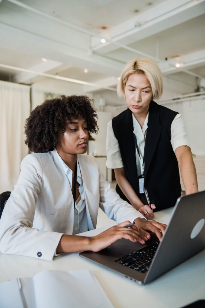 Two females looking at a open silver laptop working on something. Both have pens in their hands and look focused. The female on the left has curly hair, a grey blazer and is typing. The female on the right has straight above the shoulders hair with a white shirt and black vest blazer. These two women could be working for their apprenticeship.