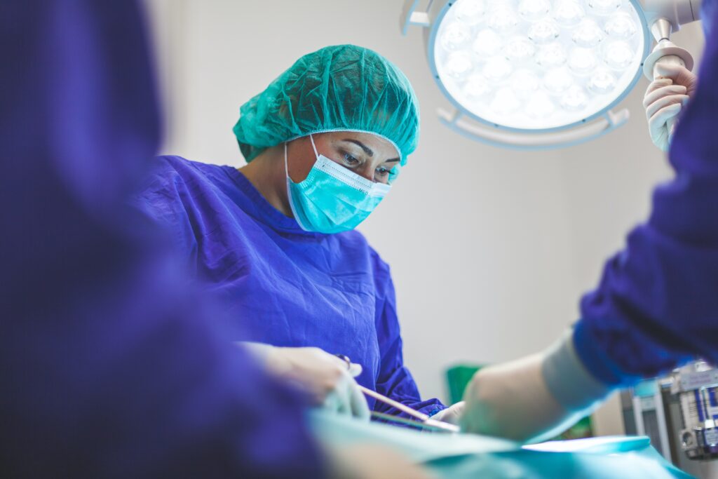 A medical professional in blue scrubs, gloves, and hairnet in an operating theatre with other colleagues assisting them.