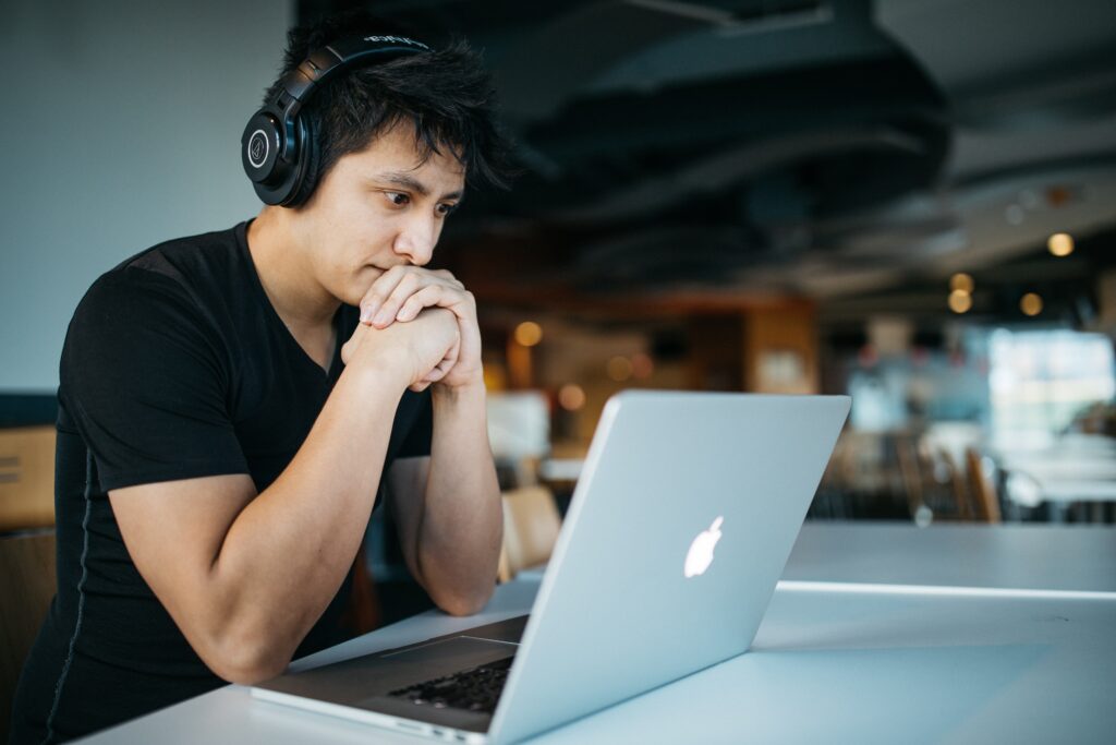 A male presenting person with dark hair sits at a desk with arms up to his face, hands in a fist. The person is wearing headphones and is concentrating on a laptop.